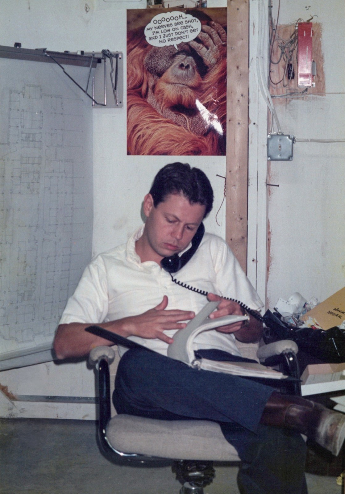 A young Allen on the phone in a job-site trailer, blueprints and a wry poster behind him